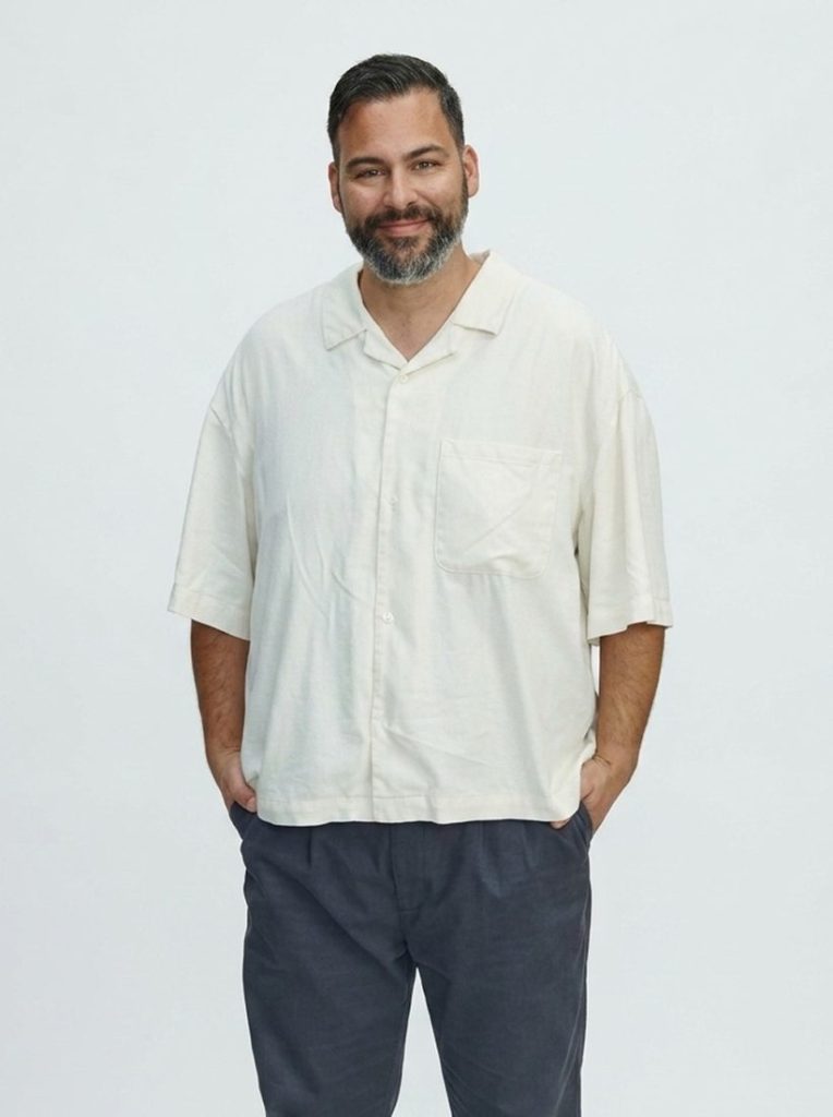 A smiling man with a grey beard in a cream shirt, hands in his pockets